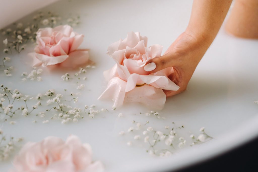 Hand einer Frau mit weißem Nagellack hat in einem milchfarbenen Becken mit Rosenblüten eine Rose in der Hand